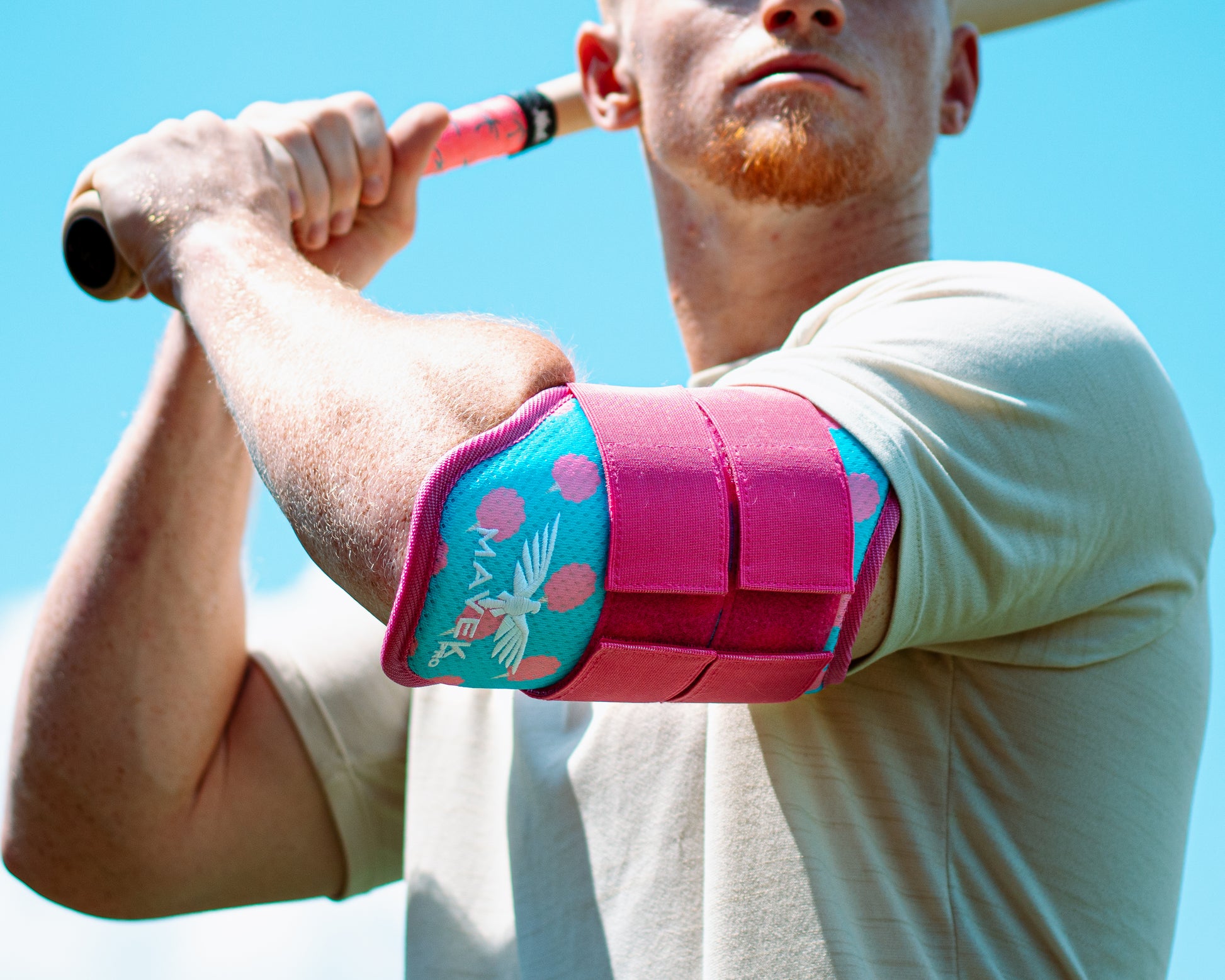 Man holding a baseball bat with a pink and blue elbow guard on his arm against a clear blue sky.