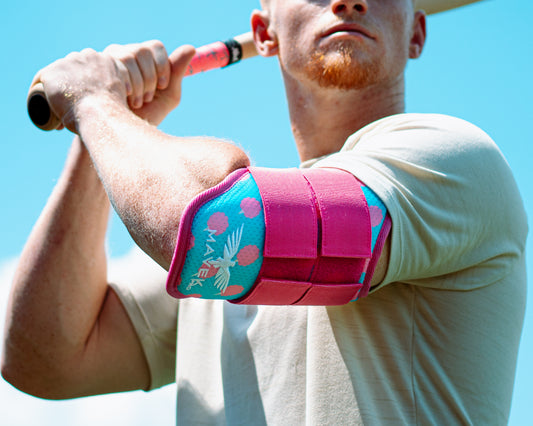 Man holding a baseball bat with a pink and blue elbow guard on his arm against a clear blue sky.